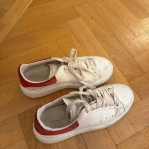 Alexander McQueen vita sneakers röd detalj - Säljer ett par ikoniska Alexander McQueen sneakers i vitt skinn med chunky sula och röda mockadetaljer på hälen. Klassisk rund tå, snörning och diskret logga bak. Perfekta för dig som vill ha en clean men ändå statement look.