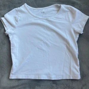Vit croppad t-shirt från H&M - En basic vit croppad t-shirt från H&M med rund halsringning och korta ärmar. Perfekt att matcha med jeans eller kjol för en clean och enkel look. Gjord i mjuk bomull som känns skön mot huden.