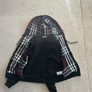 Svart hoodie från Burberry - Svart hoodie från Burberry med klassiskt rutig insida i huva och foder, dragkedja framtill och ribbade muddar. Ikonisk design med snygga detaljer i rött, vitt och grått. Perfekt för dig som vill ha en stilren och lyxig streetwear-look.