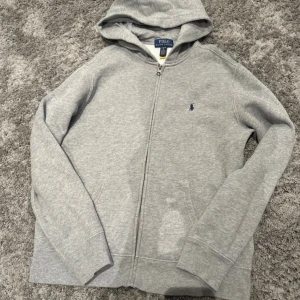Grå hoodie från Polo Ralph Lauren - Snygg grå hoodie från Polo Ralph Lauren med dragkedja framtill och klassisk broderad logga på bröstet. Tröjan har huva, känguruficka Perfekt för chill dagar eller när du vill ha en clean look. Säljer även därför ingen användning Nypris 1200kr stl (164)