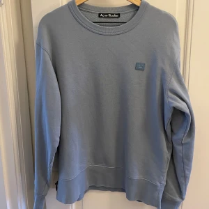 Ljusblå sweatshirt från Acne Studios - Säljer en stilren ljusblå sweatshirt från Acne Studios med klassisk rund hals och det ikoniska Face-loggan broderad på bröstet. Tröjan är i mjuk bomull och har en relaxed passform med långa ärmar. Perfekt för en clean och avslappnad look.
