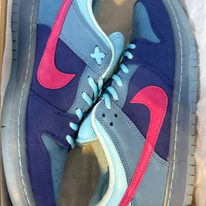 Nike Dunk SB dunk low run the jewels - Nike SB Dunk Low Run The Jewels är en iögonfallande sneaker med en fet mix av ljusblå och mörkblå mocka, kombinerat med en stark rosa Swoosh. På hälen finns ett guldigt Nike-broderi och under skon en genomskinlig blå sula med textdetaljer. Skorna har rund tå, platt sula och klassisk snörning, och levereras med tre olika skosnören i olika nyanser av blått, så du kan anpassa stilen precis som du vill.  Skorna är helt nya och aldrig använda.