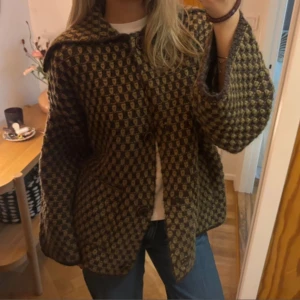 Brun rutig oversize kappa från Zara - Säljer denna populära bruna och beige rutig kappa från Zara med oversized passform, stora knappar och bred krage. Jackan är stickad i ullblandning och har vida ärmar samt två stora fickor framtill. Perfekt för dig som gillar retrovibbar och statement-plagg. Helt slutsåld. Använd en gång. 