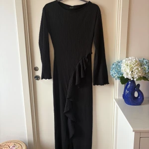 Svart långklänning från Gina Tricot - Svart långklänning från Gina Tricot i strukturerat tyg med långa ärmar och volangdetalj framtill. Endast använd en gång. Klänningen har en figurnära passform och lätt utsvängda ärmslut för en cool look. Perfekt till: ✨ Middagar eller dejtkvällar 🎭 Teater- eller konsertbesök 🥂 Födelsedagsfiranden och cocktailfester 💃 Höst- och vinterfester där du vill vara elegant men bekväm