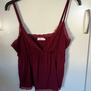 Vinröd linnetopp med spets från Nelly - Supersöt vinröd linnetopp från Nelly med tunna axelband och spetsdetaljer längs urringning och nederkant. Toppen har en liten rosett framtill och är lätt utställd för en avslappnad look. Perfekt att styla med jeans eller kjol.