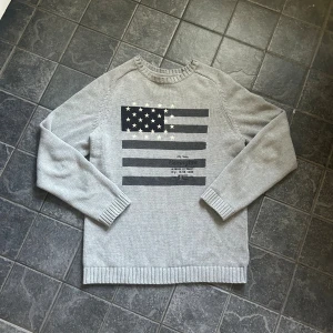 Grå stickad tröja Lexington M - Snygg grå stickad tröja från Lexington med amerikansk flagga framtill i svart och vitt. Tröjan har rund hals, ribbade muddar och är långärmad. Perfekt för dig som gillar klassisk och avslappnad stil.