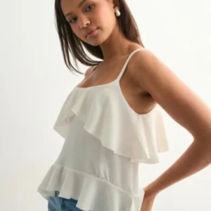 Vit volangtopp från Nelly - Säljer en superfin vit linnetopp från Nelly med tunna axelband och dubbla volanger både upptill och nertill. Toppen har en luftig känsla och är perfekt till jeans eller kjol. Materialet är lätt strukturerat och ger en snygg look.