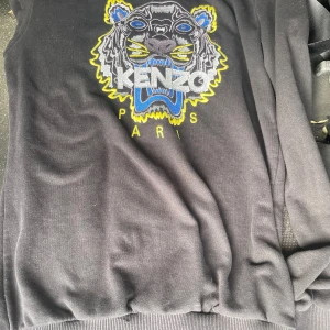 Svart Kenzo tröja med tigerbrodyr - Svart tröja från Kenzo med ikonisk tigerbrodyr i blått, gult och grått framtill. Rund halsringning och långa ärmar. Tröjan är i mjuk bomull och har ribbade muddar vid ärmslut och nederkant. Perfekt statement piece för dig som gillar streetstyle. Pris kan diskuteras!