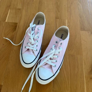 Rosa Converse  - Säljer ett par klassiska Converse sneakers i ljusrosa canvas med vita snören och vit gummisula. Skorna har rund tå och svart rand längs sulan. Perfekta för dig som vill ha en stilren och fräsch look med en touch av färg.