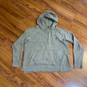 Grå hoodie från Polo Ralph Lauren - Snygg grå hoodie från Polo Ralph Lauren med klassisk huva och känguruficka. Tröjan har en liten mörkblå logga broderad på bröstet och är tillverkad i mjuk bomullsmix. Perfekt för chill dagar eller när du vill ha en clean och enkel look.