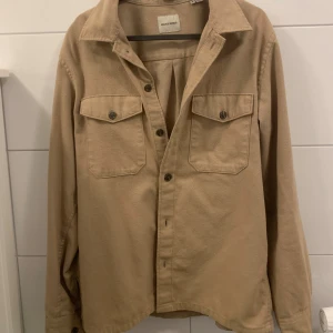 Beige overshirt från Jack & Jones - Stilren beige overshirt från Jack & Jones med klassisk krage, två bröstfickor med lock och knappar framtill. Skjortan är i mjuk bomull och har långa ärmar med manschett. Perfekt lager-på-lager-plagg för en chill look.
