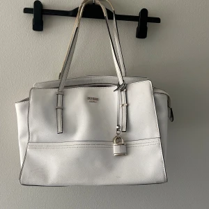 Vit handväska från Guess med hänglås - Snygg vit handväska från Guess med stilrena linjer och silverfärgat hänglås som detalj. Väskan har två långa handtag, dragkedja upptill och en praktisk ficka på baksidan. 