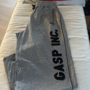 Grå mjukisbyxor från GASP XL - Snygga grå mjukisbyxor från GASP i storlek XL. Byxorna har resår och snörning i midjan, dragkedjeficka fram och stor bakficka med GASP INC.-tryck. Perfekta för chill eller gymmet, tillverkade i mjukt bomullsmaterial.
