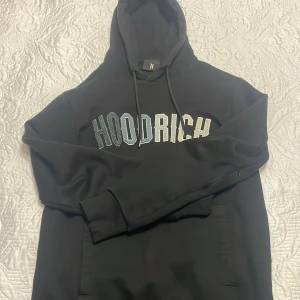 Svart hoodie från Hoodrich XS - Svart hoodie från Hoodrich i storlek XS med stor broderad logga i vitt och grått över bröstet. Klassisk huva med snörning och långa ärmar. Perfekt för dig som gillar streetwear och vill ha en stilren look.