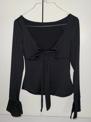 Svart långärmad topp med öppning och knyt - Svart långärmad topp med v-ringning och snygg öppning och knytning framtill. Ärmarna har en liten slits och materialet känns mjukt och stretchigt, troligen polyester. Modellen är figurnära och croppad, perfekt för att ge en cool och trendig vibe.