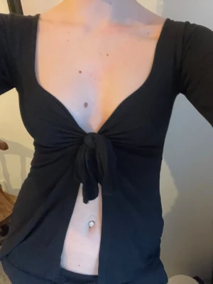 Svart croppad knytblus - Säljer en svart croppad blus med djup urringning och knyt framtill. Toppen har lång ärm och är tillverkad i mjukt, stretchigt material som sitter snyggt på kroppen. Perfekt för dig som gillar trendiga och lite vågade plagg.