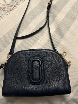  Ny Mörkblå axelväska från Marc Jacobs - Snygg mörkblå axelväska från Marc Jacobs i äkta skinn med justerbar rem och guldfärgade metalldetaljer. Väskan har en rundad form, dekorativ tofs och det klassiska Marc Jacobs-logot på framsidan. Perfekt storlek för det viktigaste och stilren design.