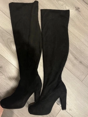Svarta höga stövlar med klack - Säljer ett par svarta, overknee stövlar med hög klack och plattform. Snygg, smal siluett och rund tå. Tillverkade i mjuk mocka som ger en lyxig känsla. Perfekta för att lyfta din outfit och ge extra längd.