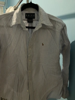 Randig skjorta från Ralph Lauren - Klassisk skjorta från Ralph Lauren i blåvitt randigt mönster. Skjortan har lång ärm, knappar framtill och den ikoniska broderade loggan på bröstet. Tillverkad i 100% bomull för en skön och luftig känsla.