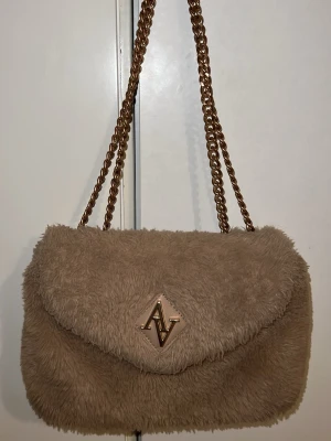 Beige fluffig axelväska från AV - Säljer en beige fluffig axelväska från AV med guldfärgad kedja och magnetlås. Väskan har ett mjukt pälsliknande material på utsidan och flera fack inuti, inklusive ett med dragkedja. Perfekt för dig som gillar mysiga och trendiga accessoarer. Dragkedjan är dock lite rostat!