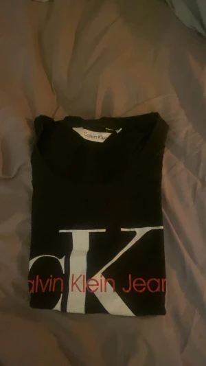 Svart Calvin Klein Jeans t-shirt - Svart t-shirt från Calvin Klein Jeans med stor vit CK-logga och röd text på framsidan. Klassisk rund hals och korta ärmar. Perfekt för dig som gillar stilrena märkesplagg med tydlig branding.