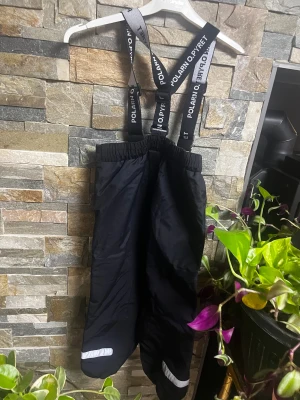 Svarta täckbyxor Polarn O. Pyret 110 - Svarta täckbyxor från Polarn O. Pyret med justerbara hängslen och reflexdetaljer på benen. Byxorna har elastisk midja och är perfekta för kalla dagar. Materialet är slitstarkt och vattenavvisande syntet. Nmr 1