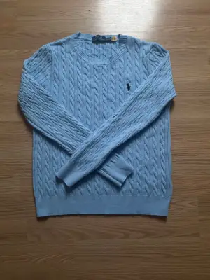 Snygg ljusblå stickad tröja från Polo Ralph Lauren med klassiskt kabelstickat mönster och broderad logga på bröstet. Rund halsringning och långa ärmar. Perfekt för dig som gillar stilrena och tidlösa plagg.