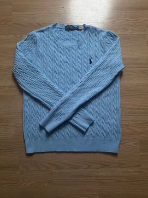 Ljusblå stickad tröja Polo Ralph Lauren - Snygg ljusblå stickad tröja från Polo Ralph Lauren med klassiskt kabelstickat mönster och broderad logga på bröstet. Rund halsringning och långa ärmar. Perfekt för dig som gillar stilrena och tidlösa plagg.