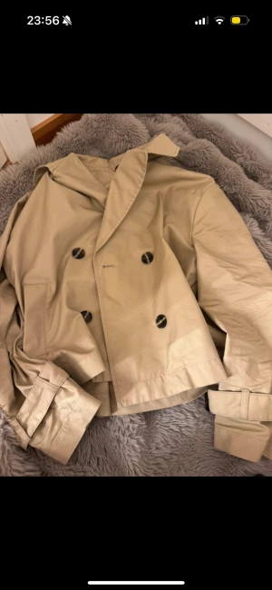 Beige trenchcoat ny pris 779 - Snygg beige trenchcoat med dubbelknäppning och klassisk krage. Jackan har bälte vid ärmsluten och stora svarta knappar framtill. Perfekt för dig som gillar stilrena och tidlösa plagg med lite edge.