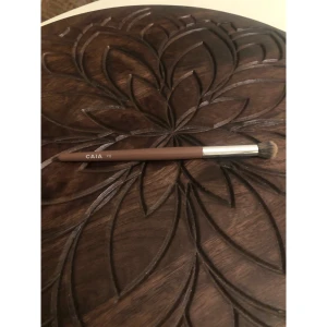 CAIA F12 sminkborste  - Säljer en CAIA F12 ögonskuggsborste med brunt skaft och silverfärgad metallhylsa. Borsten har mjuka, täta strån i beige och svart som är perfekta för att placera concealer. Stilren design som gör sminkrutinen extra lyxig.