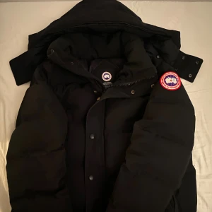 Svart dunjacka från Canada Goose - Säljer en svart dunjacka från Canada Goose med ikonisk röd och vit patch på ärmen. Jackan har huva, dragkedja och tryckknappar framtill. Perfekt för kalla vinterdagar och har en riktigt schysst puffig look. Passar dig som vill ha både stil och värme.