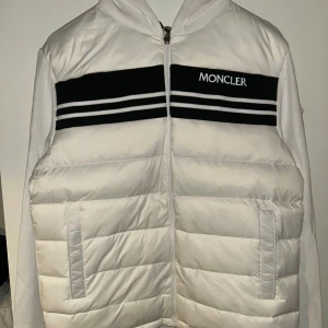 Vit pufferjacka med huva från Moncler - Säljer en snygg vit pufferjacka från Moncler med huva och dragkedja. Jackan har svarta och vita ränder över bröstet samt Moncler-logga. Framsidan är quiltad och har två fickor. Ärmar och rygg är i mjukt tyg för extra komfort.