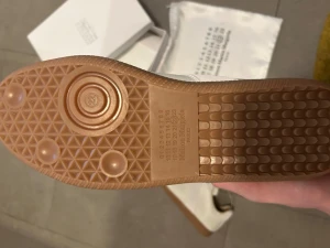 Maison Margiela vita sneakers - Maison Margiela sneakers i ljusbeige och vitt med klassisk gummisula i brunt. Snygg mix av skinn och mocka på ovandelen, rund tå och platt sula. Diskret snörning och stilren design som ger en clean vibe. Perfekt för dig som gillar minimalistiskt mode.