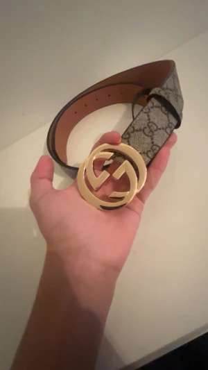  bälte med guldspänne - Snyggt  bälte med klassiskt monogrammönster i beige och brunt. Bältet har ett stort, rundat guldspänne med dubbla G-loggan. Tillverkat i skinn och canvas, perfekt för att lyfta vilken outfit som helst.