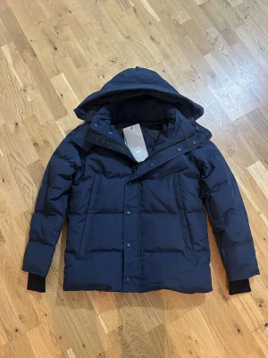 Mörkblå dunjacka Canada Goose M - Säljer en mörkblå dunjacka från Canada Goose i storlek M. Jackan har huva, dragkedja och knappar framtill, samt ribbade muddar vid ärmslut. Fylld med dun och har en clean, quiltad look. Perfekt för kalla dagar och har snygg passform.