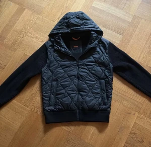 Svart quiltad hoodiejacka från BOSS - Svart cardigan från BOSS med quiltad front och huva i syntetmaterial, samt stickade ärmar och rygg. Jackan har dragkedja framtill och ribbade muddar. BOSS-logga på vänster ärm ger en clean och exklusiv vibe. Pris kan diskuteras ? Om du har några frågor är de bara att höra av dig.