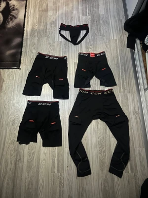 Hockeyutrustning – Hockeystavar och byxor - En samling av damask hockeyutrustning bestående av tre par damask hockeybyxor och ett par hockeykalsonger. Utrustningen är från varumärket CCM och är utformad för att ge optimalt skydd och komfort för damhockeyspelare. Byxorna är slitstarka, bekväma och har praktiska fickor för enkel förvaring, perfekt för både träning och match.
