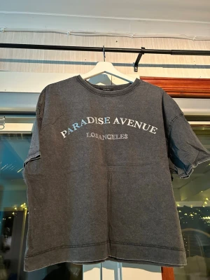 Grå t-shirt strl. M - En fin t-shirt som tyvärr blev lite använd. ”Paradise avenue” tryck med rhinestones under (alla kvar). En liten slit längst ner på båda sidorna som lägger till en fin detalj. Inte mycket använd.