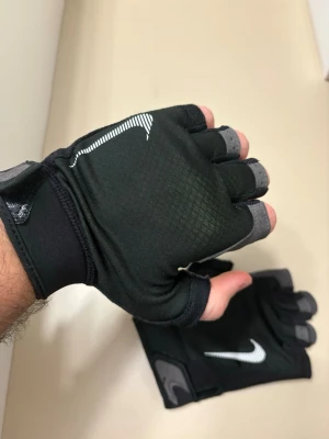 Nike  träningshandskar - Svarta fingerlösa träningshandskar från Nike med grå vadderade insidor och vit logga på ovansidan. Snygg design med justerbar kardborre vid handleden och ventilerande material för extra komfort under gympasset. Aldrig använt 