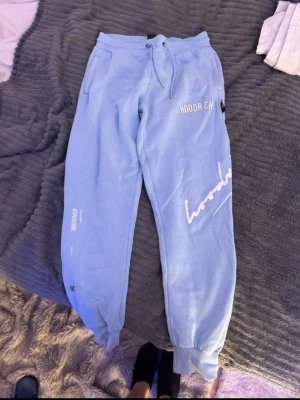 Blå mjukisbyxor från Hoodrich - Snygga ljusblå mjukisbyxor från Hoodrich med vita tryckdetaljer och logga på benet. Byxorna har snörning i midjan, muddar vid bensluten och sidofickor. Perfekta för en chill och avslappnad stil. Den har en pytteliten fläck, men det märks inte alls av