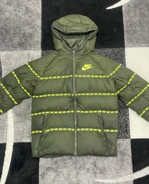 Olivgrön pufferjacka från Nike XL - Säljer en olivgrön pufferjacka från Nike i storlek XL. Jackan har huva, hel dragkedja och coola gula Nike-loggor längs ärmar och bröst. Perfekt för kalla dagar och riktigt snygg streetstil. Standard fit och tillverkad i syntetmaterial.