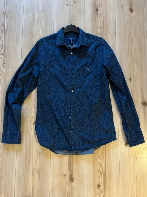 Blå blommig skjorta från Gant - strl.M - Snygg blå skjorta från Gant med mörkblått blommigt mönster. Klassisk krage, knäppning framtill och diskret bröstficka med Gant-logga. Skjortan är i bomull och har långa ärmar. Perfekt för dig som vill ha en unik twist på din outfit.