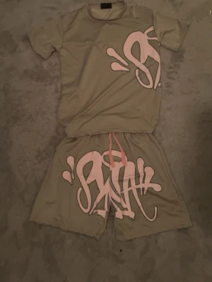 Beige shorts med rosa graffiti-tryck - Säljer ett par beige shorts med stort rosa graffiti-inspirerat tryck framtill. Shortsen har snörning i midjan och är tillverkade i ett mjukt, syntetiskt material som känns skönt mot huden. Perfekta för dig som gillar streetwear och vill sticka ut.