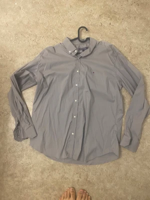 Grå skjorta från Tommy Hilfiger XXL - Klassisk grå skjorta från Tommy Hilfiger i storlek XXL. Skjortan har lång ärm, knappar framtill och liten broderad logga på bröstet. Snygg och enkel design i bomull, perfekt till jeans eller chinos.