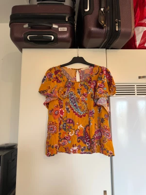 Blommig blus från Odd Molly - Säljer en färgstark blus med blommigt och paisleyinspirerat mönster i orange, lila, blått och rött. Blusen har korta ärmar, rund hals och knapp i nacken. Luftigt material som känns skönt mot huden, perfekt för vår och sommar. Från märket Odd Molly. Storlek 3 som enligt deras storleksguide motsvarar en L. Mycket fint skick, jag hittar inga synliga anmärkningar eller defekter. 