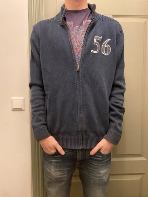 Mörkblå stickad kofta/ cardigan med dragkedja från Bondelid - Säljer en riktigt snygg mörkblå stickad kofta från Bondelid med broderad siffra 56 på bröstet. Koftan har ribbad krage, muddar och nederkant samt praktisk dragkedja framtill. Riktigt fet både som overshirt eller som tjocktröja. 