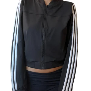 Svart Adidas Neo ziptröja med ränder - Svart ziptröja från Adidas Neo med klassiska vita ränder längs ärmarna och diskret logga på ryggen. Tröjan har dragkedja framtill, ribbade muddar och en sportig, croppad passform 🖤