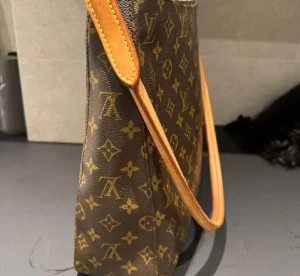 Louis Vuitton brun axelväska med monogram - Säljer en klassisk brun axelväska från Louis Vuitton med ikoniskt monogrammönster i guld. Väskan har en stilren rektangulär form, dragkedja upptill och ljusbrunt skinnhandtag. Insidan är ljus och har en praktisk ficka. Perfekt för dig som gillar lyxiga detaljer.