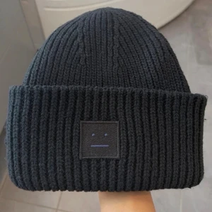 Svart stickad mössa Acne Studios - Svart ribbstickad mössa från Acne Studios med klassisk face-logga framtill. Tillverkad i mjuk ull och har en bred uppvikt kant. Perfekt för att hålla värmen och ge en clean look under kalla dagar.