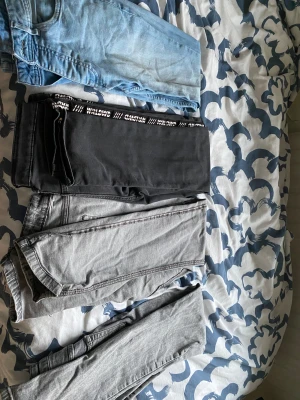 Tre par jeans och ett par svarta byxor - Säljer tre par jeans i olika tvättar: ett par ljusblå jeans, ett par grå jeans och ett par mörkgrå jeans. Dessutom ett par svarta byxor med vit textdetalj längs sidan. Alla har klassisk femficksdesign och raka ben, perfekta för en chill och stilren look.